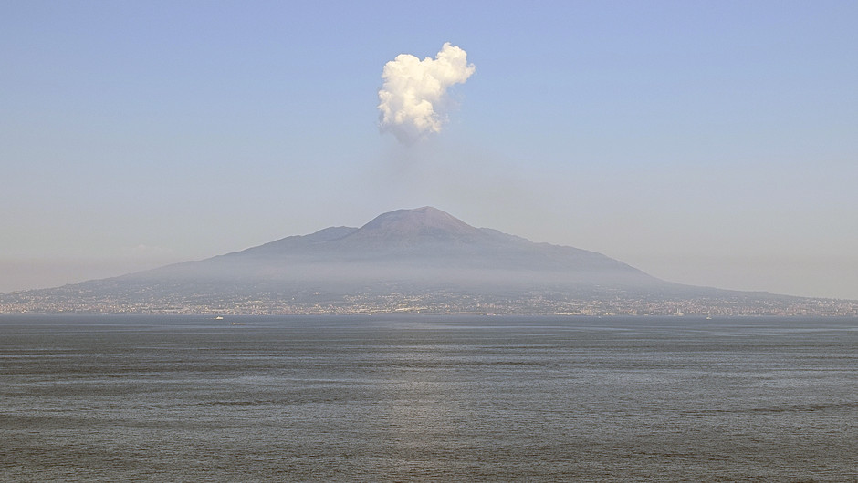 Vulkanen Vesuvius sedd från håll. Staden med samma namn ligger strax nedanför, i förgrunden hav. Ovanför vulkanen syns ett vitt moln.