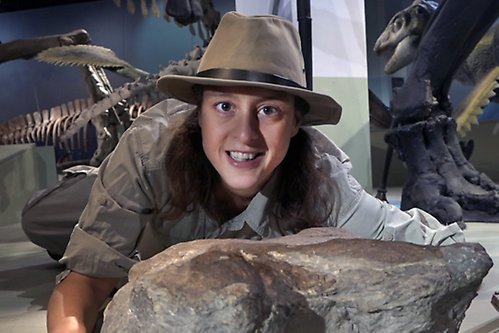 Dino-Doris i hatt i utställningen omgiven av dinosaurier och fossil.