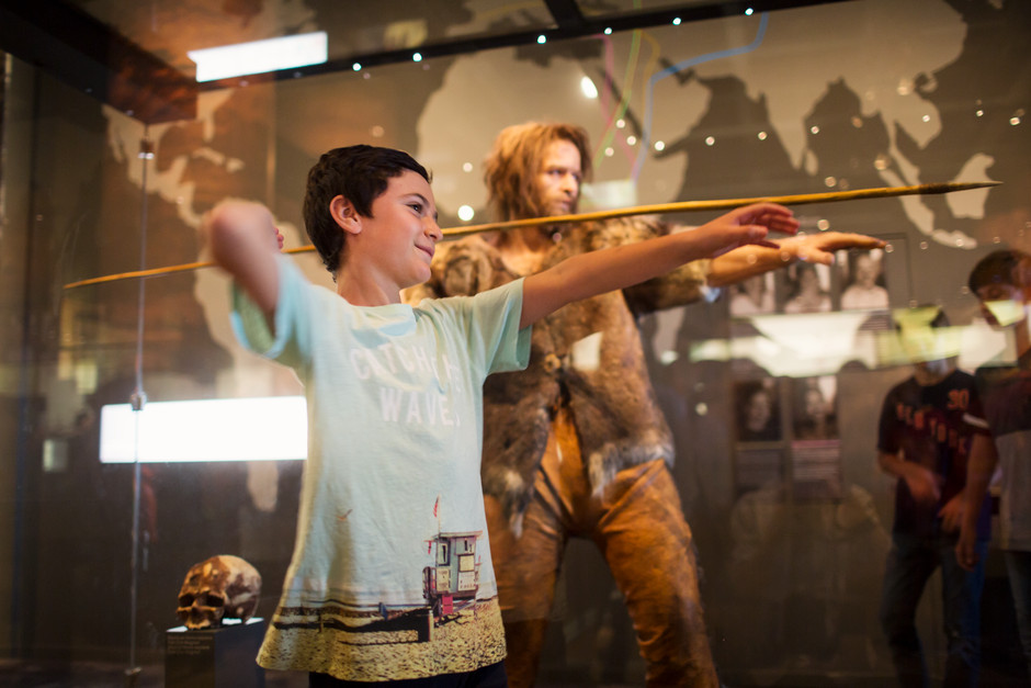 "Photo of a boy pretending to throw a spear in front of an exhibition display where a prehistoric person is throwing a spear.