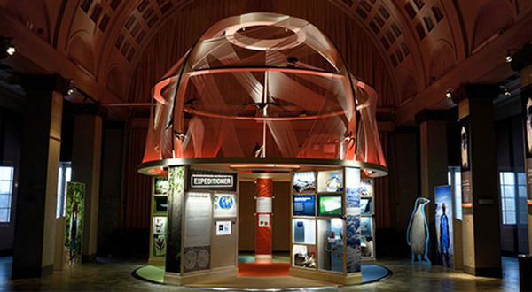A small, brightly-lit exhibition about scientific expeditions with many photos, viewed from the outside, featuring a rounded roof.